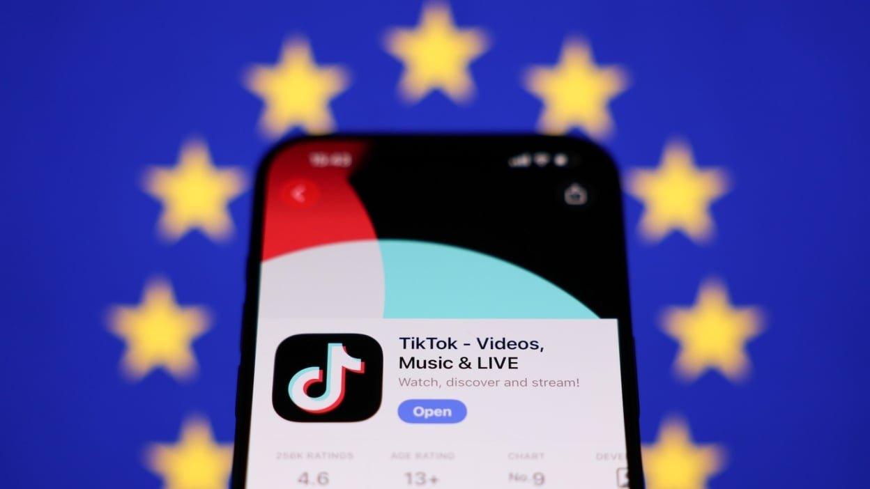 A phone showing TikTok in an app store is held in front of the EU flag.