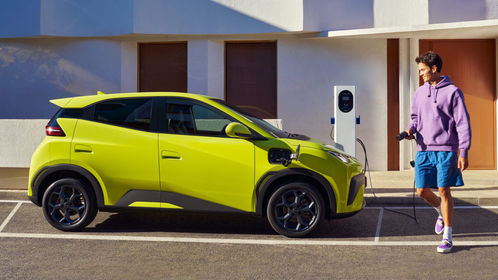 The BYD Atto 1 in lime green next to a charger. A man holding the charging cord walks up to it to plug it in.